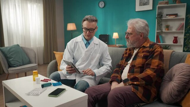Full-size video of a doctor checking up the condition of his patient at home. He is using tablet to explain something to an elderly man with health issues.