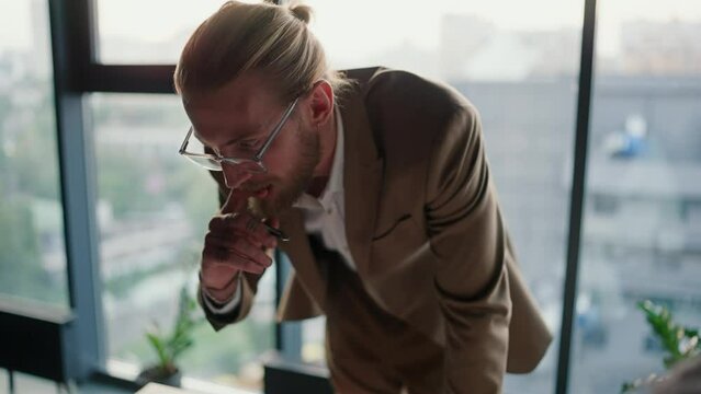 Close-up shot of a blond man in glasses with a beard in a light suit leans his hands on the table and tells his colleagues what needs to be done in a modern office