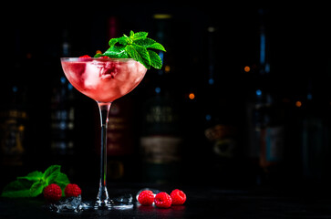 Raspberry daiquiri pink cocktail with white rum, sugar syrup, fruit juice, lime, berry and ice in glass. Black bar counter background, bar tools and bottles