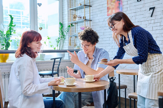 Woman in an apron with cup of coffee and plate of cake serving young people in cafeteria
