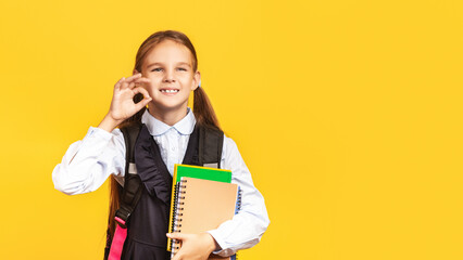 Back To School. Cute Schoolgirl Gesturing OK Holding Books Posing Over Yellow Studio Background. Copy Space