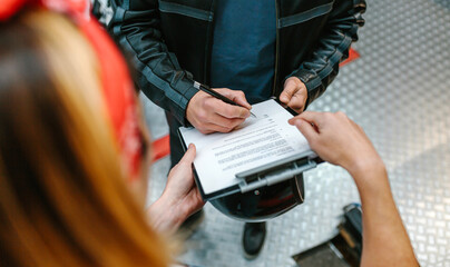 Unrecognizable female mechanic with red hair bandana holding a clipboard while biker man wearing leather jacket signing insurance policy to receipt his repaired motorcycle