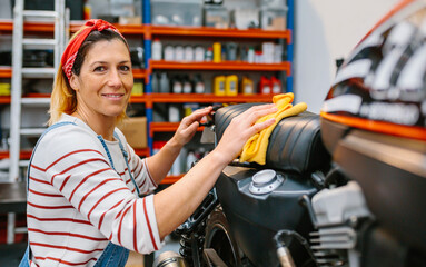 Fototapeta na wymiar Portrait of smiling female mechanic with freckles and hair bandana looking at camera while cleaning motorcycle seat with a microfiber cloth on factory
