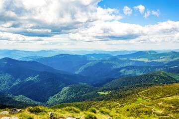 Naklejka na ściany i meble Landscape in green meadows and blooming flowers and snow-capped mountain. Mountains ridge high rocky peaks. Mountain landscape with hiking trail. Mountain path. Springtime landscape in mountains