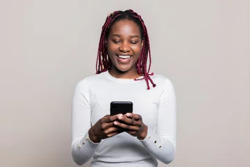 Fototapeten Happy dreamy delighted African American dark skinned woman with colourful braids standing in the studio isolated holding cellphone in hands checking messages sms chatting with friends online © ABCreative