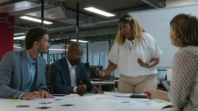 Multiracial group of coworkers in businesswear talking and gesturing by whiteboard during meeting