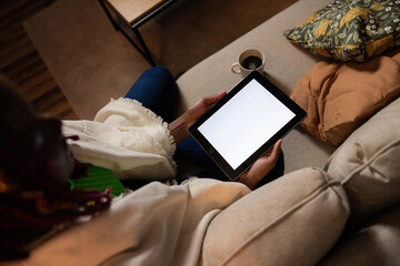 Top view of afro american dark skinned woman sitting n the sofa holding tablet with white screen drinking coffee reading latest news in the internet online evening time.