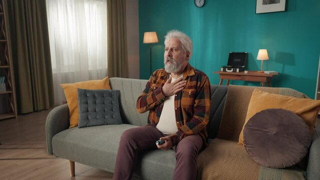 Zoom in video of an eldery, retired man, senior citizen sitting on a couch, sofa, coughing and breathing heavily, using an inhaler to ease the struggle.