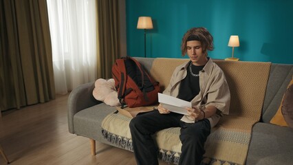 Shot of a teenager, young man sitting on a sofa, couch, reading a document, paper, admission,...