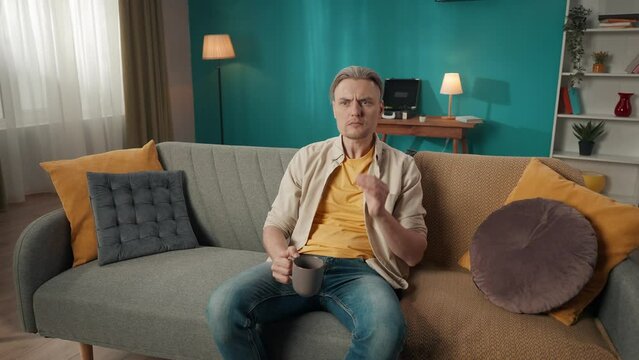 Zoom in video of a middle-aged man sitting on a couch, sofa, drinking tea, when suddenly he feels a strong toothache, pain and touches his cheeck.