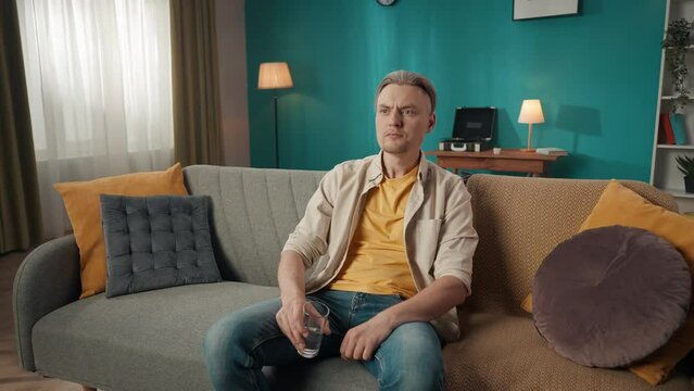 Zoom in video of a middle-aged man sitting on a couch, sofa, drinking water, when suddenly he feels a strong toothache, pain and touches his cheeck.