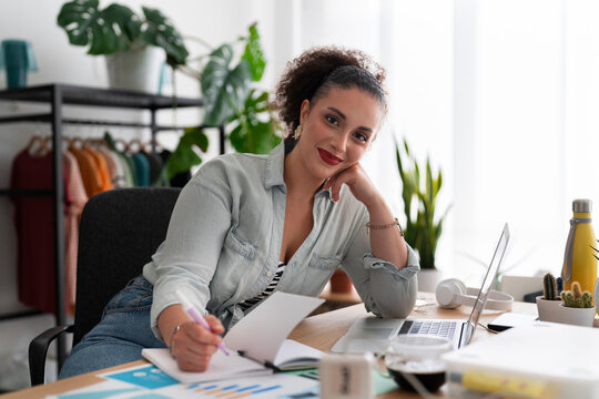 Portrait of optimistic Middle Eastern female seamstress with computer looking at camera and smiling while writing in organizer and working in showroom