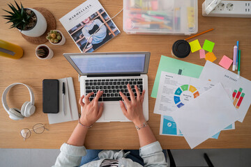 Top view of crop anonymous female freelancer with portable computer and papers analyzing data and working on remote project at desk
