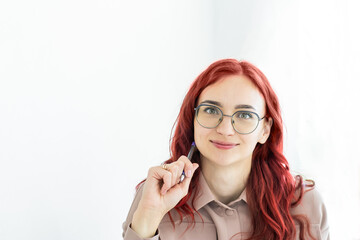 A student girl with glasses with bright hair close-up in a light dress looks and smiles while holding a pen in her hands. Study, online work, office, secretary