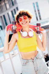 Young beautiful smiling hipster woman in trendy summer clothes. Carefree woman with curls hairstyle, posing in the street. Positive model outdoors. Cheerful and happy. Holds penny skateboard