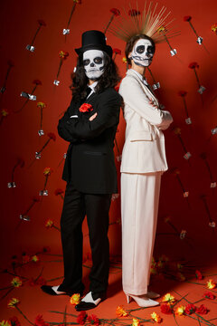 trendy couple in catrina makeup standing back to back with folded arms on red backdrop with flowers