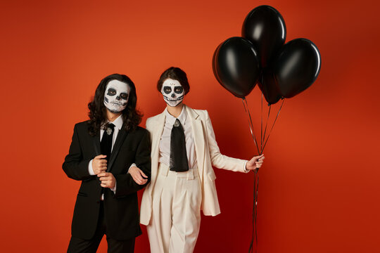 dia de los muertos party, elegant couple in sugar skull makeup standing with black balloons on red