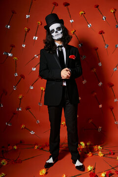 full length of man in skull makeup and festive attire on red floral backdrop, dia de los muertos