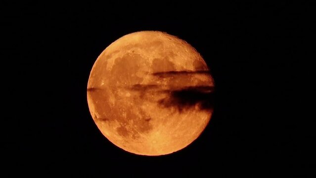 The full moon rises in the dark night sky in close-up, black clouds fly by. Real shooting through a telephoto lens