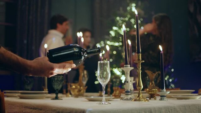 In front of the camera capturing video glass of sparkling wine on the Christmas dinner table someone pour the sparkling one on the the glass on the background group of people celebrating the Christmas