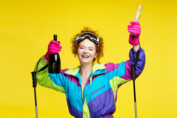 Portrait of smiling beautiful curly haired woman, skier wearing winter ski googles, holding bottle champagne or vine looking at camera isolated on yellow background. Travel, resort, vacation concept - obrazy, fototapety, plakaty