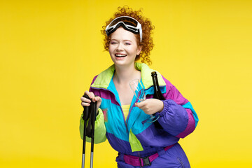 Happy beautiful curly haired woman, skier wearing winter ski googles, overalls holding bottle champagne or vine and glass isolated on yellow background. Travel, resort, winter vacation concept