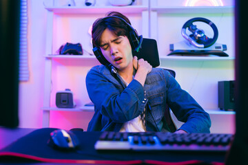 A young Asian man professional gamer wearing a jeans jacket sits on a chair and rubs a sore shoulder that suffering from stress playing a game or competition. Concept of pain from gaming and esports. © artitwpd