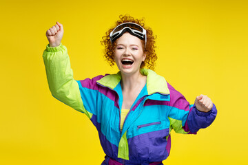 Beautiful excited curly haired woman wearing stylish overalls, ski goggles, isolated on blue yellow background. Vacation, winter, travel concept
