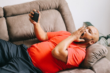 Lazy man yawns while browsing the internet or watching movies on his phone, lying lazily on the...
