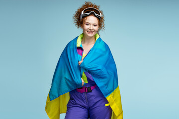 Smiling red haired beautiful woman wearing winter overalls and ski googles holding Ukrainian flag isolated on blue background - obrazy, fototapety, plakaty