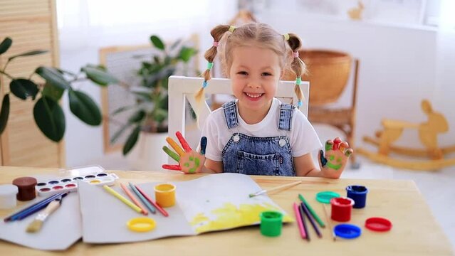 a little child girl happily makes prints in an album with her hands with a dirty face and smiles, a happy child draws a picture with gouache fingers at home at the table