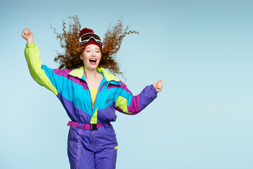 Attractive overjoyed curly haired woman wearing stylish overalls, ski goggles, winter hat jumping, having fun isolated on blue background, copy space. Vacation, winter travel concept 