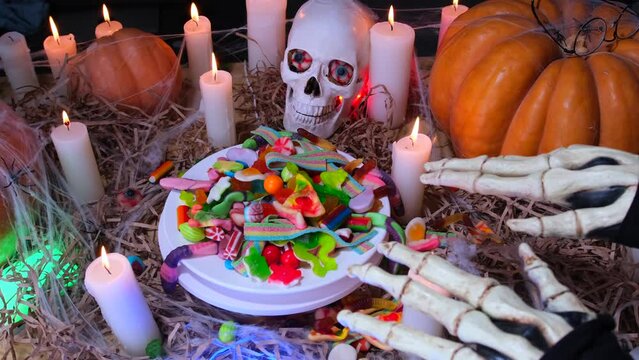 Close-up vampire hands rejoice pile colorful candies jelly sweets in crypt among candles of skulls and huge orange pumpkins in cobweb. Dramatic alluring horror scary holiday Halloween background.