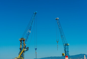 Two construction tower cranes at a construction site against a blue sky with clouds.