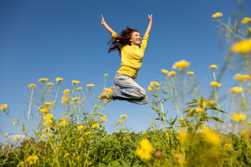 Happy and beautiful young woman in a bright yellow sweater and blue jeans  jumping high in a sunny summer field. - 651133907