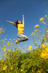 Happy and beautiful young woman in a bright yellow sweater and blue jeans  jumping high in a sunny summer field. - 651133930