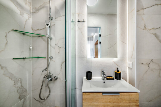 Modern tiled bathroom in white and grey cool colors. It has shower cabin with glass partition, wooden stand for white sink and mirror on the wall. Selective focus
