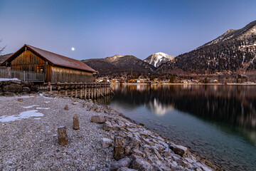 winterliche Morgenstimmung am Walchensee mit Bootshaus und Mond