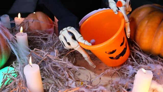 Scary funny skeleton hands demand candies in orange bucket for holiday Halloween in festive atmosphere among large pumpkins and flickering candles on cobweb. Happy Halloween party  background.