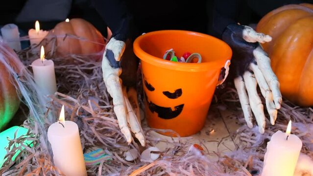 Halloween party invitation. Scary vampire hands showing many sweets in orange bucket, traditional candies spooky night Halloween celebration. Pumpkins and flickering candles.