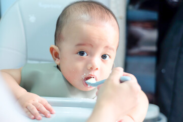 Close-up mother hand with spoon feeding a baby boy, a baby child feeding by their parent. Baby eating blend mashed food sitting on a chair, mother feeding child