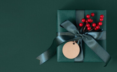 Green gift box with green ribbon, gift tag and red berries. Green background, place for your text