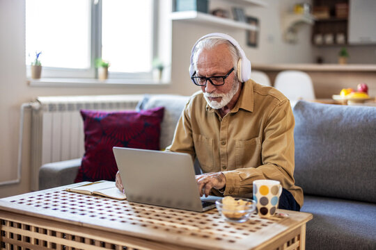 Portrait of senior man learning online with headphones and laptop at home, technology and leisure concept. Older mature man using wireless laptop apps browsing internet sit on sofa.