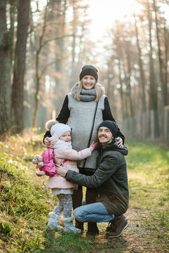 Pregnant mom, dad stand and hug daughter in background of yellow leaves in forest at sunset. Autumn family photo. Happy family walks in autumn park. Young family spending time together in nature.