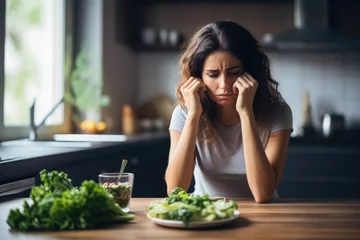 Deurstickers Woman with a plate of green salad on table in kitchen is depressed during dieting. The concept of negative consequences of a strict diet © gankevstock