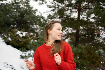 A young beautiful woman in a red sweater holds sparklers celebrating the new year against the background of snow and Christmas trees.