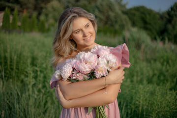 Fototapeta premium Charming natural blonde with loose hair in pink romantic dress with bouquet of peonies in nature. 