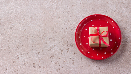 Red plate with white polka dots with a gift in craft paper tied with a red ribbon on light beige background. Simple christmas background or table
