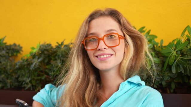 Female blogger chatting on social media using smartphone and laptop in cafe. Attractive young woman with blonde hair and glasses sitting in cafe with yellow wall background . Online communication 