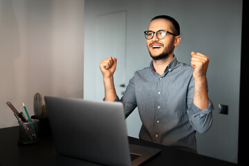 Yes. Portrait of young satisfied man working home at laptop, feels  happy about victory and work done, wearing eyeglasses and shirt. Background of grey wall.